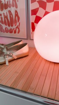 Silberne Mary Jane-Schuhe neben einer runden weißen Lampe auf einer Holzlattenoberfläche mit rot-weißer Wandkunst im Hintergrund.