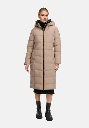 Beige lange pufferjas met een ritssluiting, capuchon en een boxy quiltontwerp. Bevat zwarte accenten en wordt gedragen met een zwarte trui en laarzen.