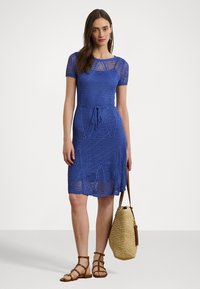 Abito blu in maglia con maniche corte, vita regolabile con cordoncino e tessuto a traforo. Abbinato a sandali marroni e una borsa di paglia.