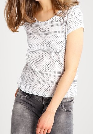 Kvinde iført en hvid kortærmet skjorte med små sorte mønstre kombineret med falmede grå jeans, stående foran en ensfarvet baggrund.