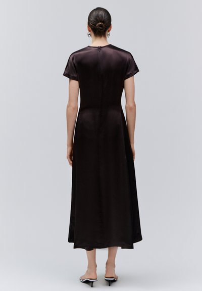 Femme avec les cheveux en chignon, portant une robe midi en satin noire à manches courtes et des mules noires à talons, debout devant un fond gris clair uni.