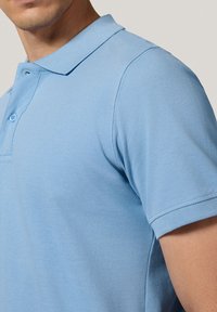 Jasnoniebieska koszulka polo z teksturowanej tkaniny, z kołnierzykiem, dwoma guzikami i krótkimi rękawami.