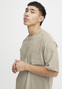 Beige bavlněné tričko s kulatým výstřihem a krátkými rukávy. Obsahuje náprsní kapsu a malou značkovou visačku na kapse. Hladká textura.