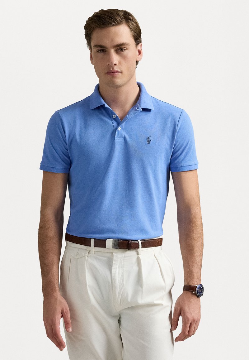 Ljusblå polo med krage och korta ärmar, som har en liten broderad logotyp. Matchas med krämfärgade byxor och ett brunt bälte.