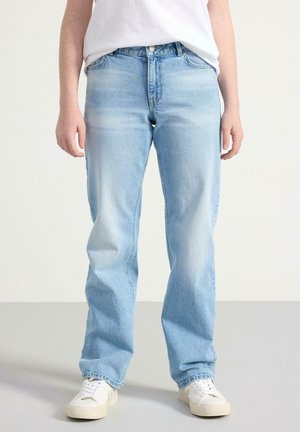 Jean en denim bleu clair avec une coupe droite, dotés d'une fermeture à bouton, de passants de ceinture et d'une texture délavée. Des baskets blanches complètent la tenue.