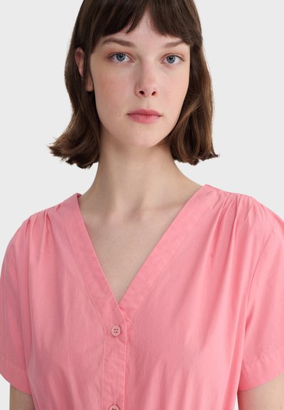 Jeune femme à la peau claire et aux cheveux bruns courts portant un chemisier rose à col en V boutonné sur un fond blanc uni.