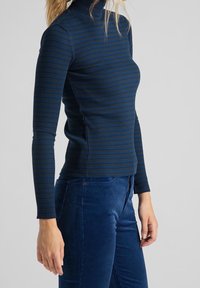 Haut à col roulé à manches longues rayé marine et sarcelle avec une texture côtelée, associé à un pantalon en velours côtelé bleu avec une fermeture éclair et une poche.