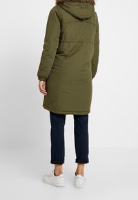 Person som bär en olivgrön parka med en furu-lined huva, marinblå uppvikta byxor och vita Reebok-sneakers, som står mot en vit bakgrund.