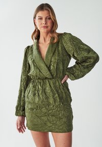 Grünes, zerknittertes Mini-Kleid mit einem tiefen V-Ausschnitt, langen Puffärmeln und einem elastischen Taillenbund. Die strukturierte Stoffqualität sorgt für visuelles Interesse.