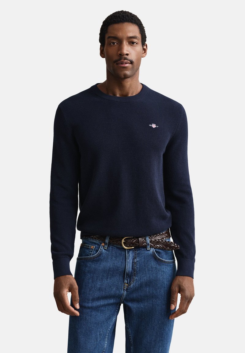 Homme portant un pull à manches longues bleu marine avec un petit emblème et un jean bleu maintenu par une ceinture tressée marron, debout devant un fond uni.