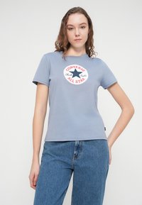 Ανοιχτό μπλε βαμβακερό t-shirt με κοντά μανίκια, με κυκλικό κόκκινο και λευκό γραφικό λογότυπο στο κέντρο. Συνδυάζεται με ψηλόμεσο μπλε τζιν παντελόνι.