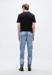 Μαύρο κοντομάνικο μπλουζάκι, ανοιχτό μπλε τζιν στενής εφαρμογής με ξεθώριασμα, κόκκινο έμβλημα με το λογότυπο HUGO στη πίσω ζώνη, στέκεται πάνω σε λείο επίπεδο.