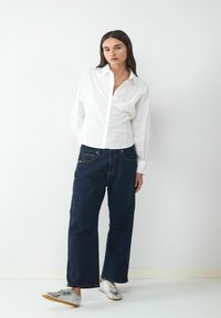 Kobieta stoi w białej koszuli zapinanej na guziki, luznych ciemnoniebieskich dżinsach i srebrnych trampkach na jednolitym białym tle.