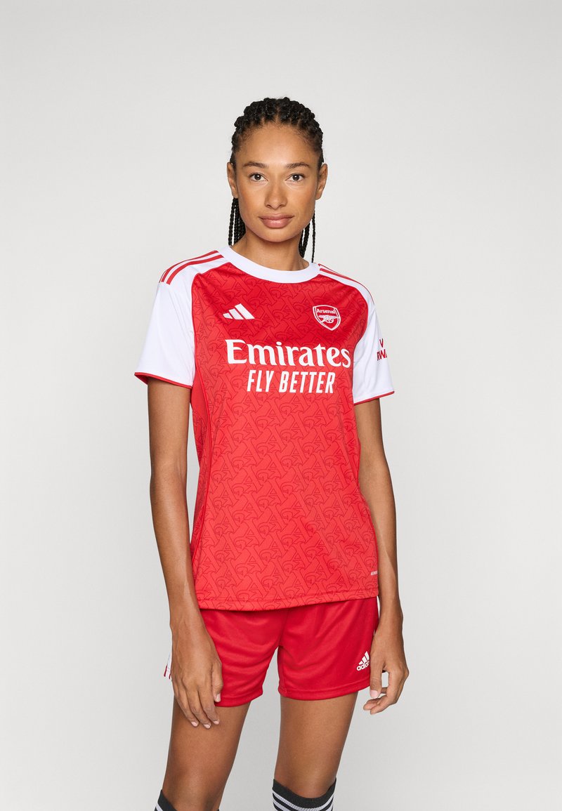 Maillot de football rouge avec des accents blancs et un logo. Présente un motif texturé, des manches courtes, et un short rouge assorti avec des rayures noires.