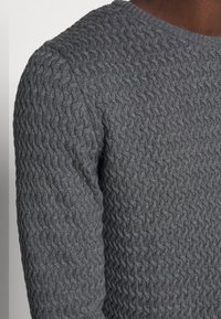 Gros plan sur une personne portant un pull en maille gris foncé texturé avec un motif torsadé en câble et un col rond.