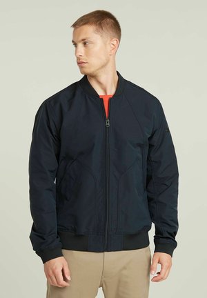 Veste de bombardier marine avec une fermeture éclair à l'avant, des poignets côtelés et des panneaux texturés, fabriquée en tissu léger. Présente un logo sur la manche.