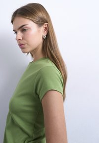 Νεαρή γυναίκα με μακριά ίσια καστανά μαλλιά, φοράει ασημένια σκουλαρίκια και πράσινο τοπ με κοντά μανίκια, μπροστά σε ένα απλό λευκό φόντο.