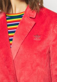 Červený kordový blejzr s širokými klopami a jemně vyšívaným logem Adidas. Pod ním se nachází tričko s barevnými pruhy, které dodává kontrast.