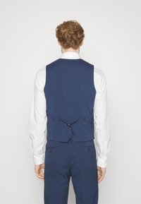 Marineblaues Weste mit tailliertem Schnitt, der bis zur Hüfte reicht, mit einem Band am Rücken. Getragen über einem weißen Langarmhemd und passenden Hosen.