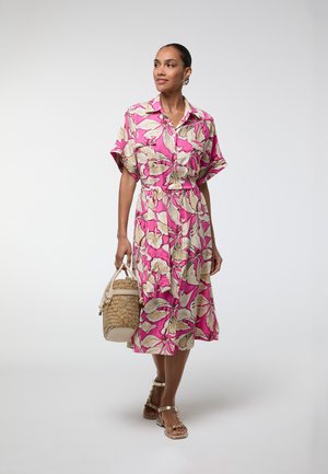 Mujer con vestido midi floral rosa y beige, sosteniendo un bolso de cesta tejido y con sandalias doradas, caminando frente a un fondo liso.