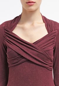 Mörkröd långärmad topp med rynkad tyg vid axlarna och en korsad design vid halsringningen. Mjukt, texturerat material.
