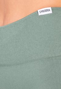 Fascia elastica a coste verde chiaro con etichetta SMILODOX piatta. Texture liscia con design aderente, ideale per abbigliamento sportivo.