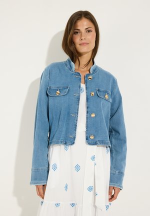 Frau trägt eine blaue Jeansjacke mit goldenen Knöpfen über einem weißen Kleid mit blauen Blumenmustern, sie steht vor einem einfarbigen hellen Hintergrund.