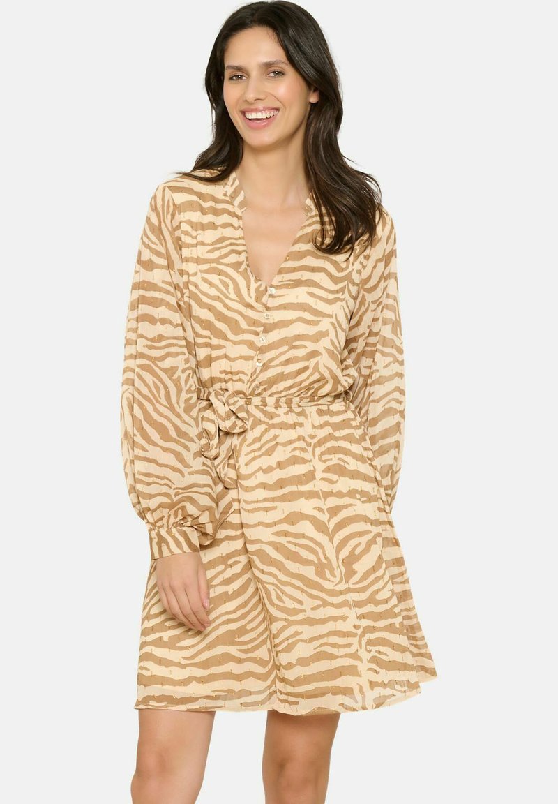 Frau mit dunklen Haaren, die lächelt, trägt ein langärmliges Kleid mit Zebra-Streifen in Beige und Hellbraun, das Knöpfe und einen Taillengürtel hat.