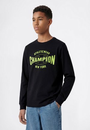 Jeune homme portant une chemise noire à manches longues avec un texte jaune "Champion Athleticwear New York" et un jean bleu, debout avec une main dans la poche.