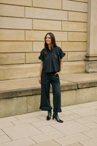 Femme debout à l'extérieur portant un chemisier noir à manches courtes volantées, un pantalon à jambes larges avec des poignets à volants et des bottines noires.