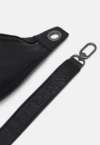 Cinghia in pelle nera con testo in rilievo "LIFESAVING" e gancio in metallo, attaccata a una borsa in pelle nera con cerniera visibile e anello in metallo.