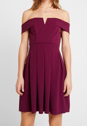 Kobieta w bezrękawowej, odkrytej na ramionach, śliwkowej sukience z dekoltem w kształcie litery V i plisowaną spódnicą, stojąca na jednolitym tle.