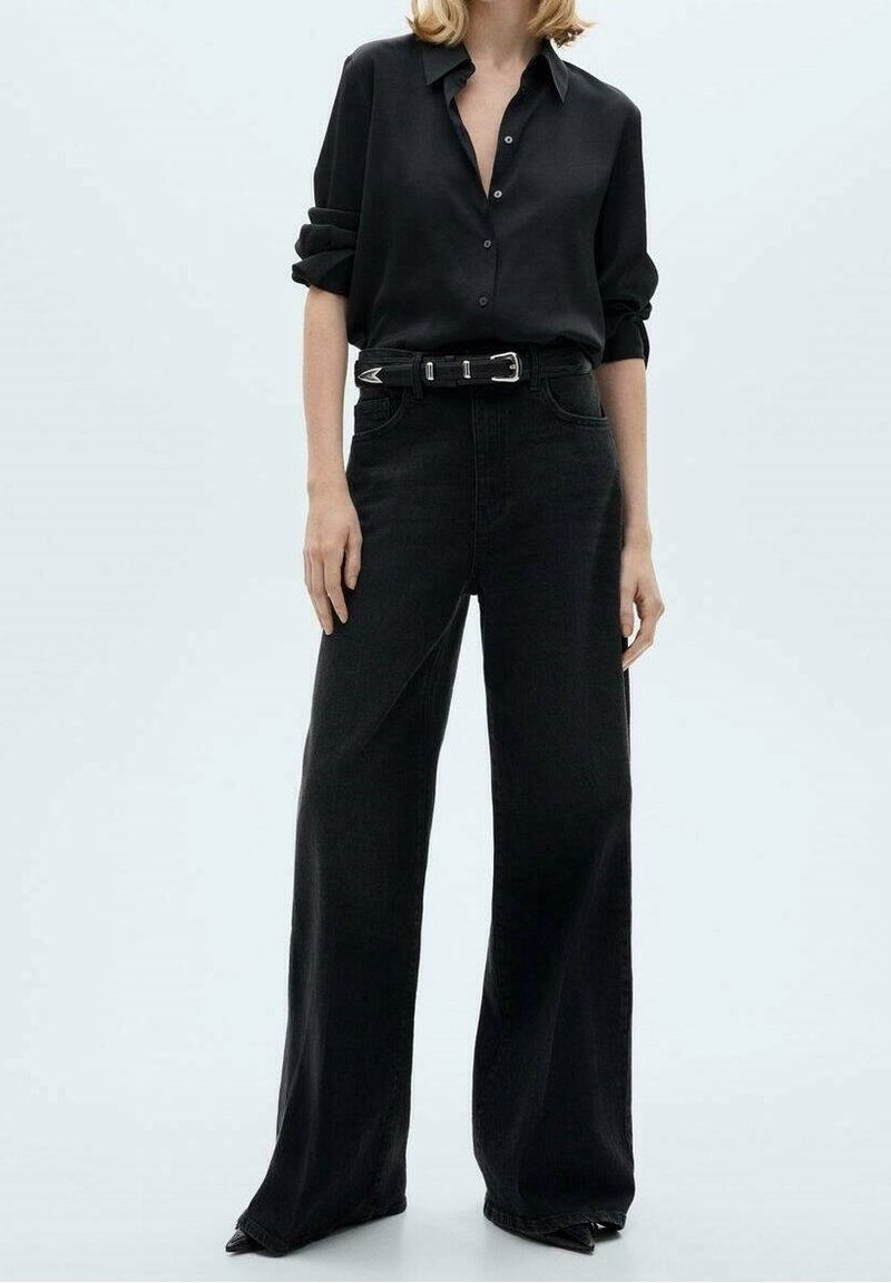 Femme portant une chemise noire à boutons avec les manches retroussées, un pantalon noir taille haute à jambes larges et une ceinture noire avec une boucle argentée.