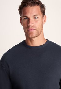 Young man with light blue eyes and short brown hair wearing a dark blue textured long-sleeve crewneck shirt, facing forward.