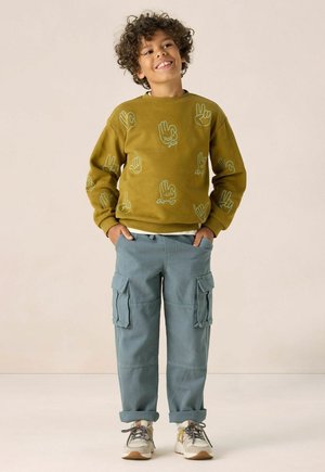 Sweatshirt graphique vert avec des motifs de mains, associé à un pantalon cargo bleu clair avec des poches latérales et des ourlets roulés. Les chaussures sont beiges et grises.