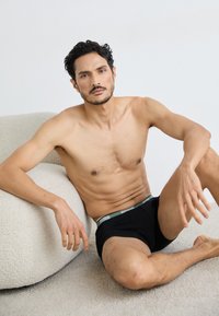 Fekete boxeralsó zöld derékkal, amelyen a márkaneve szerepel, egy texturált fehér szőnyegen ül, egy krémszínű, lekerekített kanapé mellett.