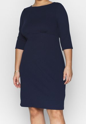 Femme portant une robe bleu marine foncé jusqu'aux genoux, à manches trois quarts et taille cintrée, debout devant un fond clair uni.
