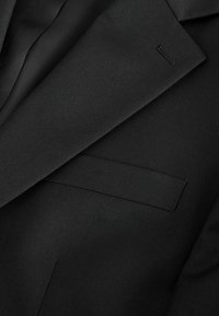 Gros plan sur le revers et la poche passepoilée d’un blazer noir, montrant la texture lisse du tissu et les détails précis des coutures.