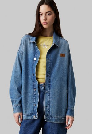 Jeansjakke i lys blå med en afslappet pasform, metalknapper og et brunt mærke. Båret over en gul ribbet top.