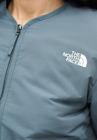 Κοντινό πλάνο ενός ατόμου που φοράει μπλε μπουφάν με φερμουάρ, με το λογότυπο "The North Face" κεντημένο στο στήθος.