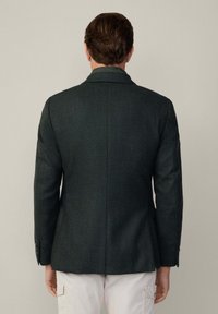 Mörkgrön texturerad blazer med en figursydd design, notchade slag och knappade ärmar, sedd från baksidan. Casual ljusa byxor.