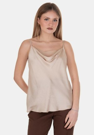 Giovane donna con capelli lunghi che indossa una canottiera di raso beige e pantaloni marroni, in piedi con un'espressione neutra su uno sfondo semplice.