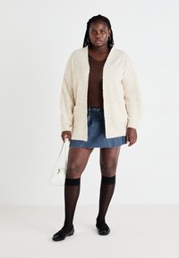 Chunky cream kleurige cardigan met een gestructureerd oppervlak, bruin aansluitend topje, spijkerrok, zwarte kniehoge kousen en zwarte platte schoenen. Witte handtas.