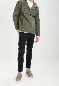 Olivgrön lätt jacka med en framficka, bärs över en vit skjorta. Kombinerad med svarta smala jeans och grågröna sneakers.