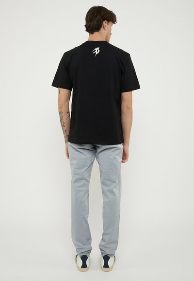 Man facing away wearing a black T-shirt with a white symbol on the upper back, light gray pants, and white sneakers with navy details.