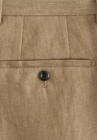 Närbild på bakficka på beige linnelångbyxor med en svart knapp och synliga sömdetaljer.