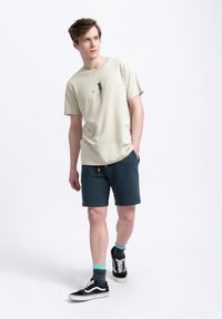 T-shirt en coton beige clair avec un petit motif sur le devant, assortie à un short bleu sarcelle foncé et des baskets noires avec des accents blancs.