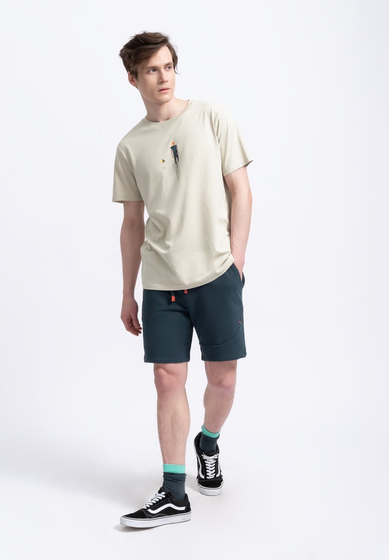 T-shirt en coton beige clair avec un petit motif sur le devant, assortie à un short bleu sarcelle foncé et des baskets noires avec des accents blancs.