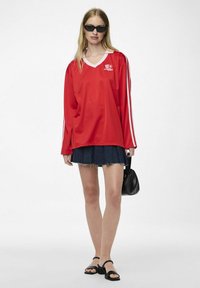 Rotes sportliches Langarmshirt mit weißem V-Ausschnitt, vertikalen weißen Streifen an den Ärmeln, kombiniert mit einem dunklen Jeansrock und schwarzen Accessoires.