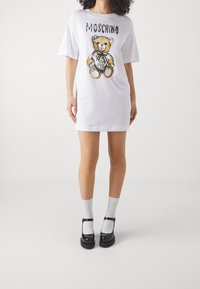 Kobieta w białej, oversize'owej sukience T-shirt z grafiką misia i napisem "Moschino", w białych skarpetkach i czarnych butach typu Mary Jane.